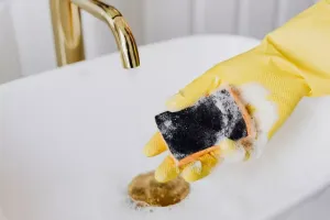 Ręka w rękawiczce czyści czarną pleśń z silikonu przy wannie za pomocą szczotki i sprayu do łazienki.